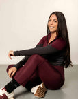 Brunette model sitting down wearing burgundy riding leggings with two tone base layer top.