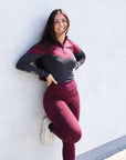 Brunette model wearing burgundy and grey base layer top with burgundy riding leggings leaning against a wall