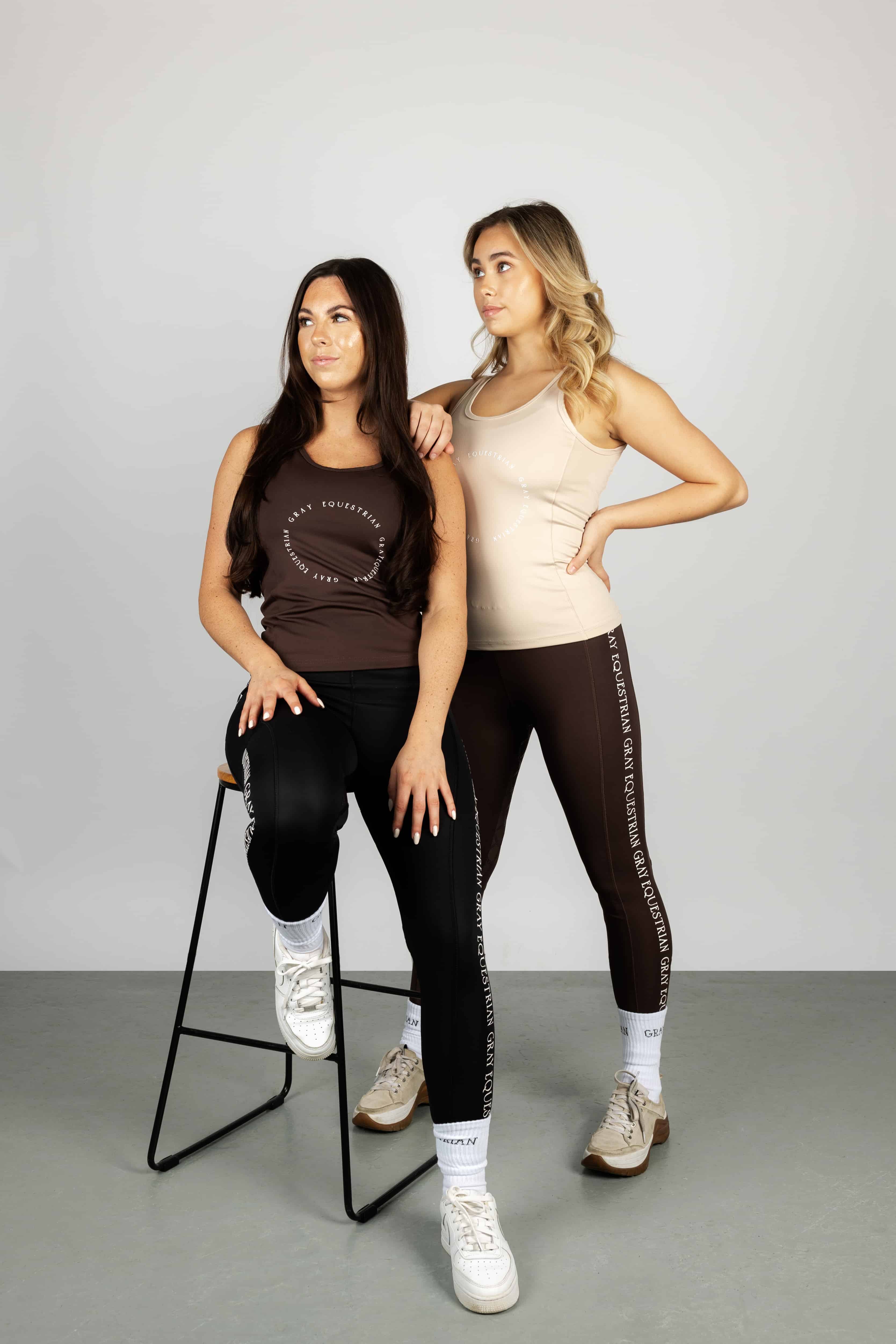Two models standing next to each other. The model on the left is wearing our brown renew vest and black riding leggings. The model on the right is wearing our nude vest and brown renew leggings.
