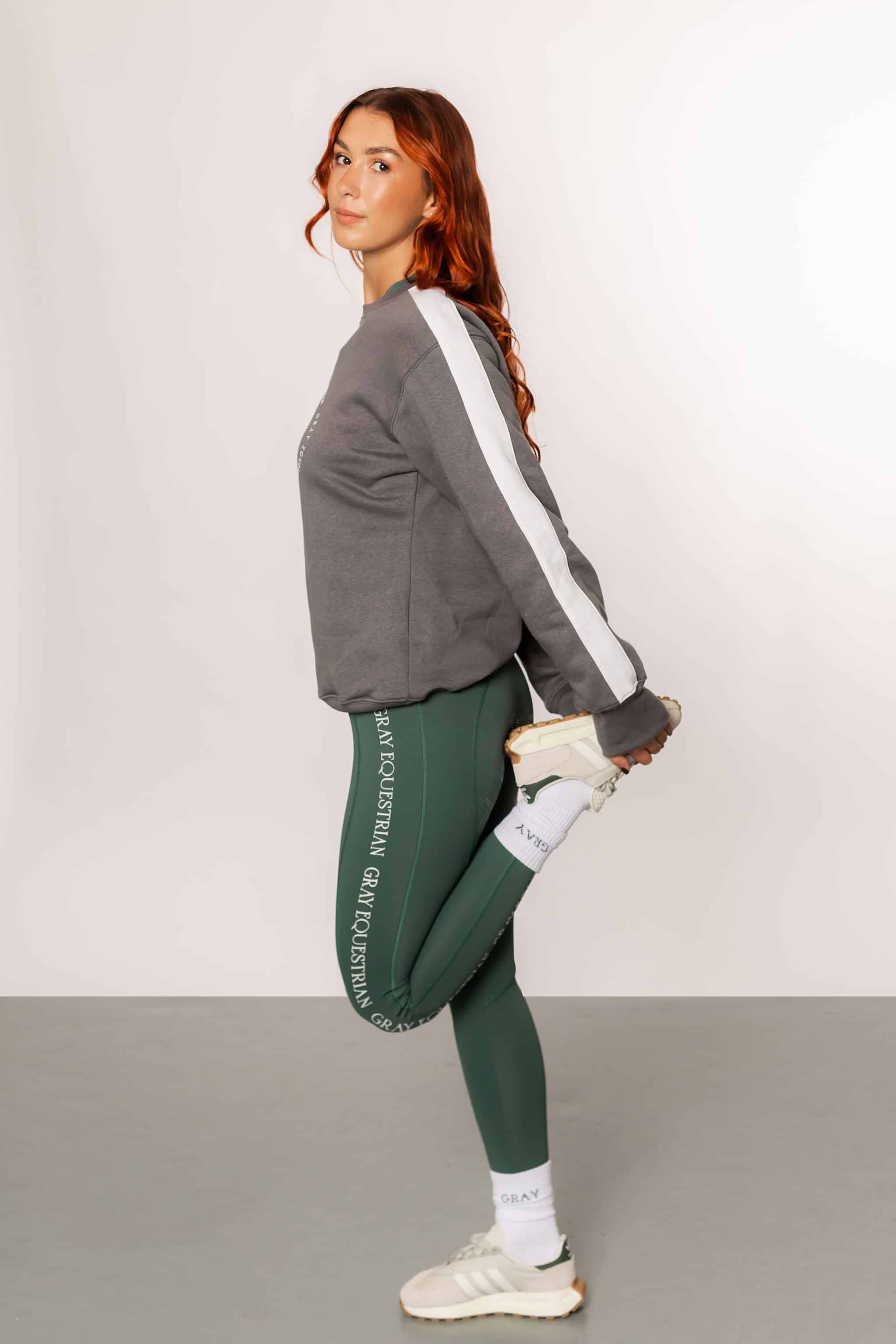 A view of the side of our grey sweatshirt with a white strip down the sleeves. The model is also wearing our green riding leggings.