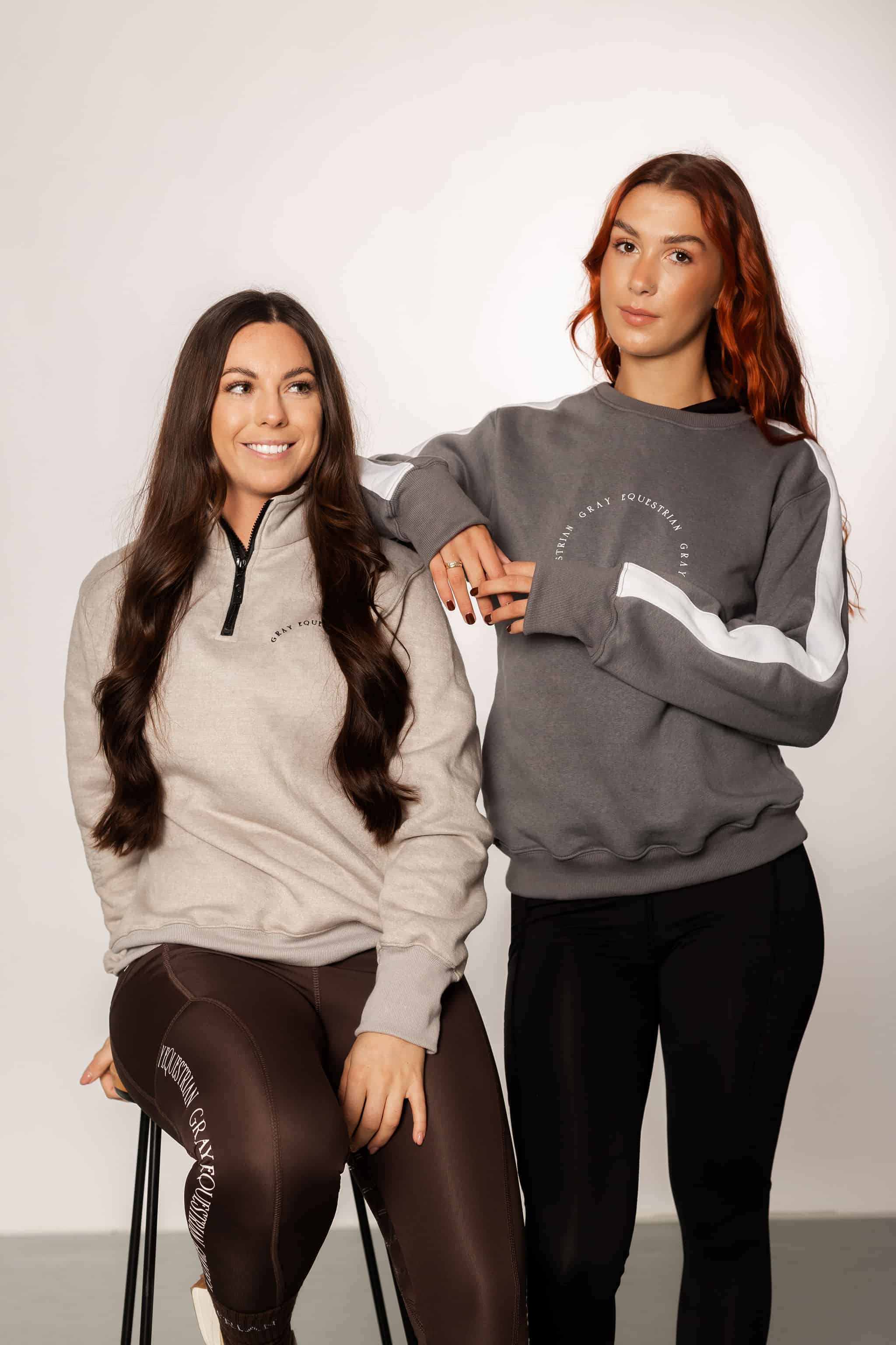 Two models standing side by side. The model on the left is wearing out taupe grey 1/4 zip sweatshirt and brown leggings. The model on the right is wearing our grey and white sweatshirt and black leggings.