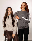 Two models standing side by side. The model on the left is wearing out taupe grey 1/4 zip sweatshirt and brown leggings. The model on the right is wearing our grey and white sweatshirt and black leggings.