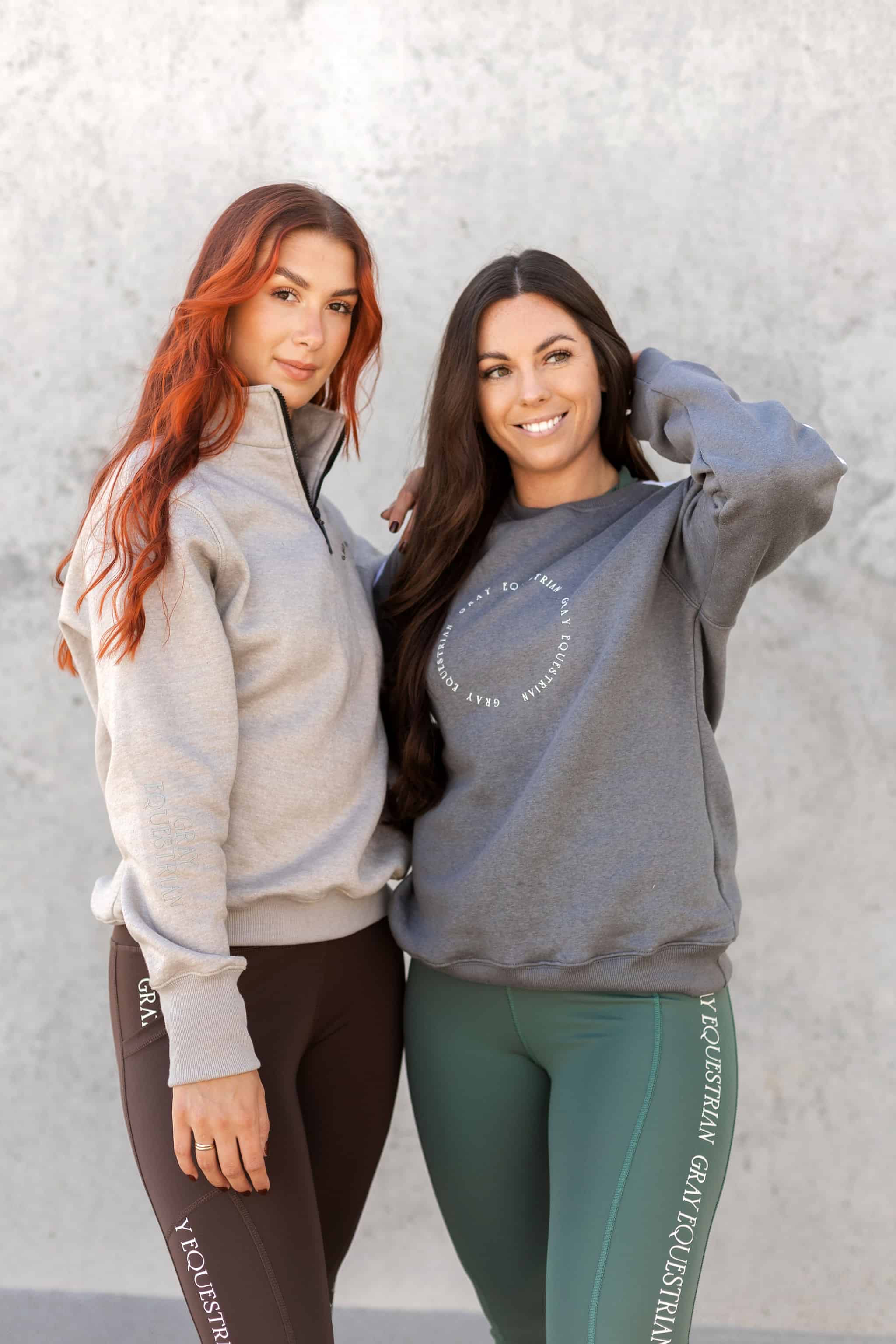 Two models standing side by side. The model on the left is wearing out taupe grey 1/4 zip sweatshirt and brown leggings. The model on the right is wearing our grey and white sweatshirt and green leggings.