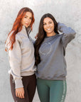 Two models standing side by side. The model on the left is wearing out taupe grey 1/4 zip sweatshirt and brown leggings. The model on the right is wearing our grey and white sweatshirt and green leggings.