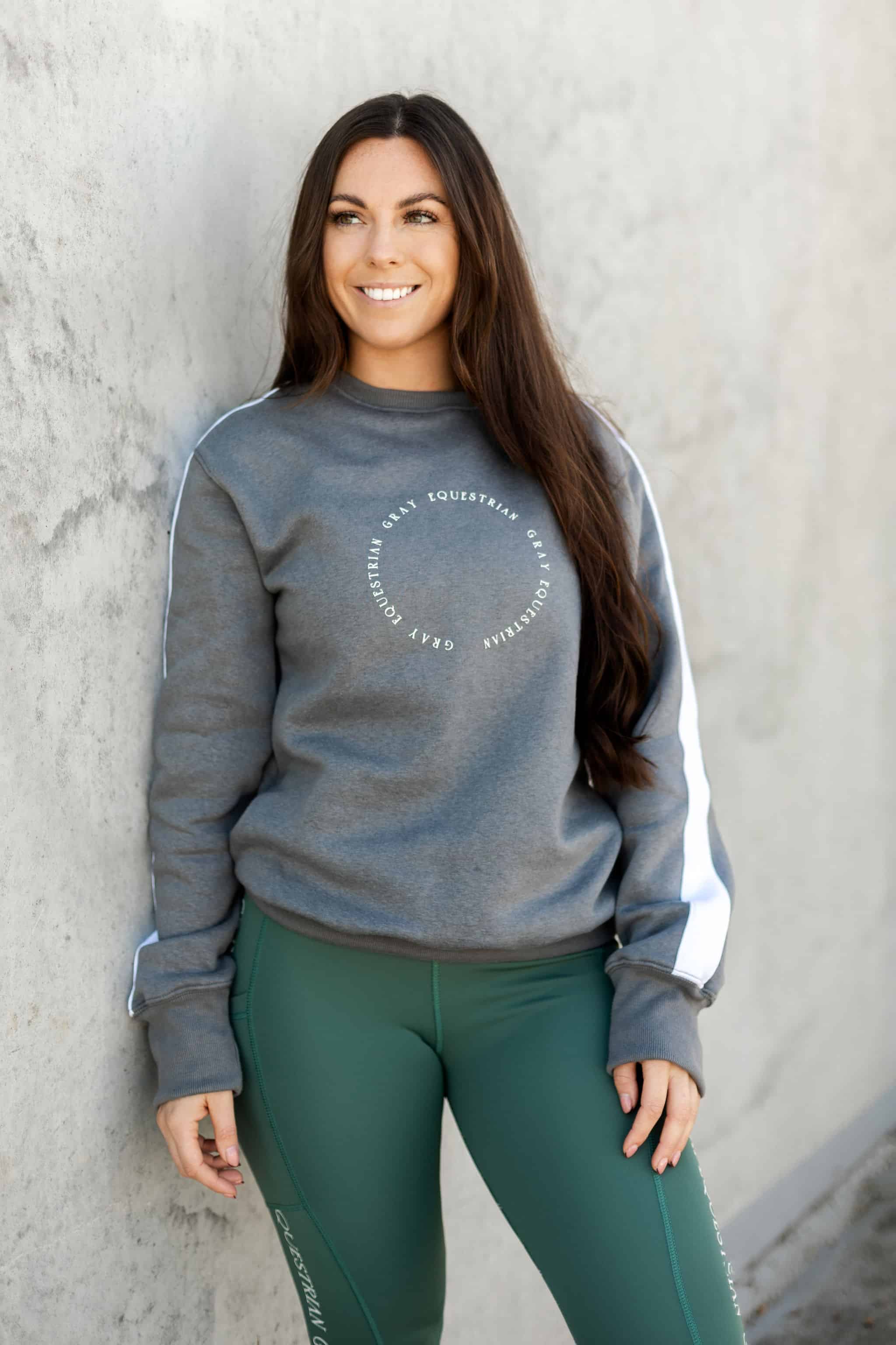 A model wearing our grey sweatshirt with white paneling down the sleeves and minimalist Grey Equestrian branding. The model is also wearing our green riding leggings.
