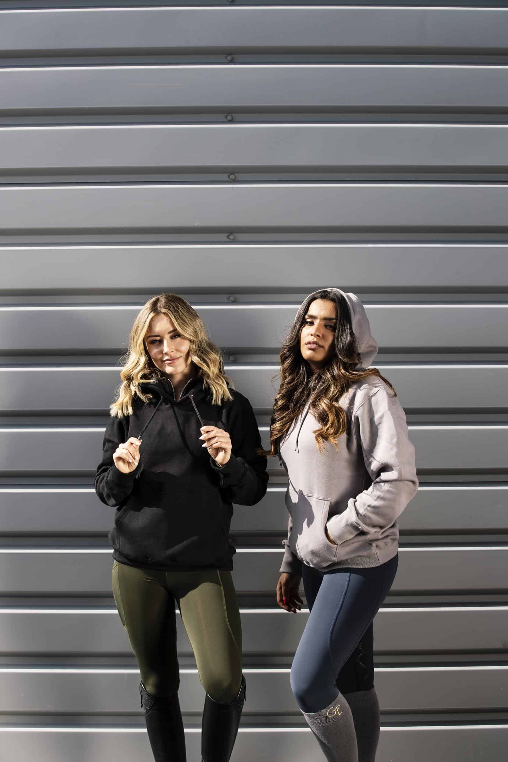 Two models standing next to each other. The model on the left is wearing our black hoodie and khaki riding shorts. The model on the right is wearing our taupe grey hoodie and navy riding leggings.