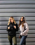 Two models standing next to each other. The model on the left is wearing our black hoodie and khaki riding shorts. The model on the right is wearing our taupe grey hoodie and navy riding leggings.