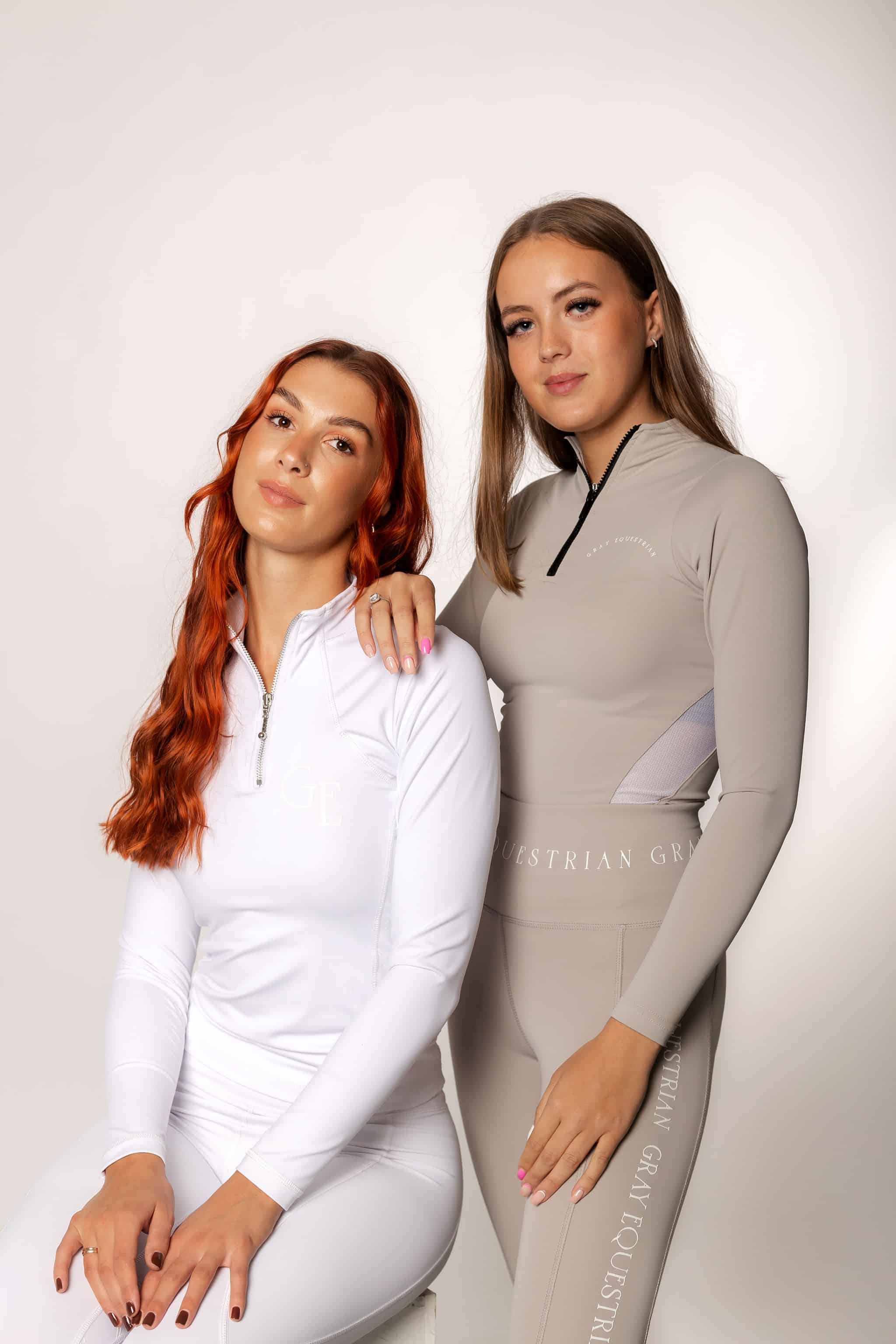 Two models next to each other. The first is sitting on a stool and is wearing our white long sleeved base layer and matching leggings. The one to the right is wearing our taupe base layer and leggings.