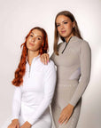 Two models next to each other. The first is sitting on a stool and is wearing our white long sleeved base layer and matching leggings. The one to the right is wearing our taupe base layer and leggings.
