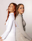 Two models sitting back to back. The one on the left is wearing our white long sleeved base layer and matching leggings. The one to the right is wearing our taupe base layer and leggings.