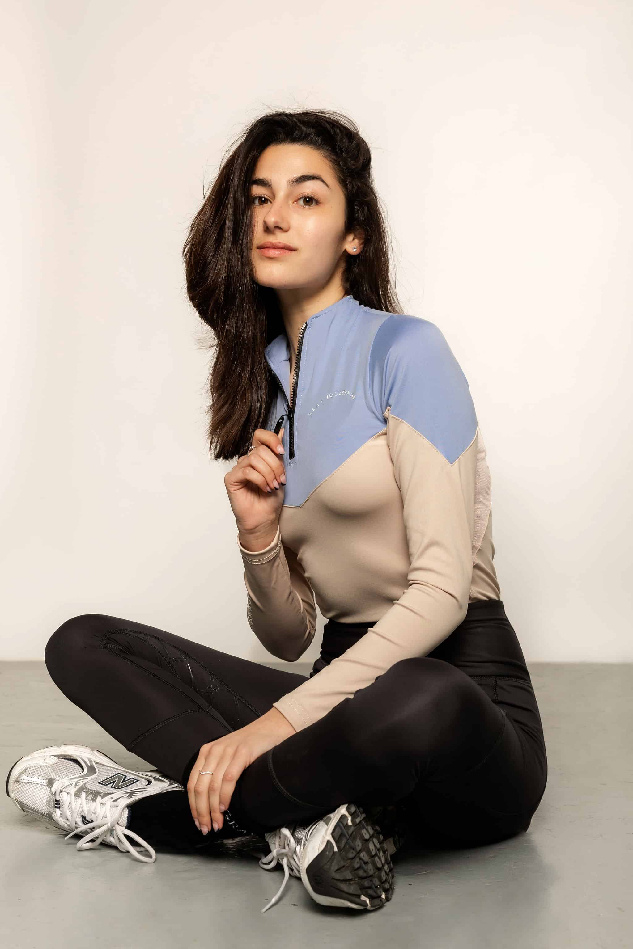 Model sitting down wearing a two tone equestrian base layer with 1/4 zip detail and mesh side panels.