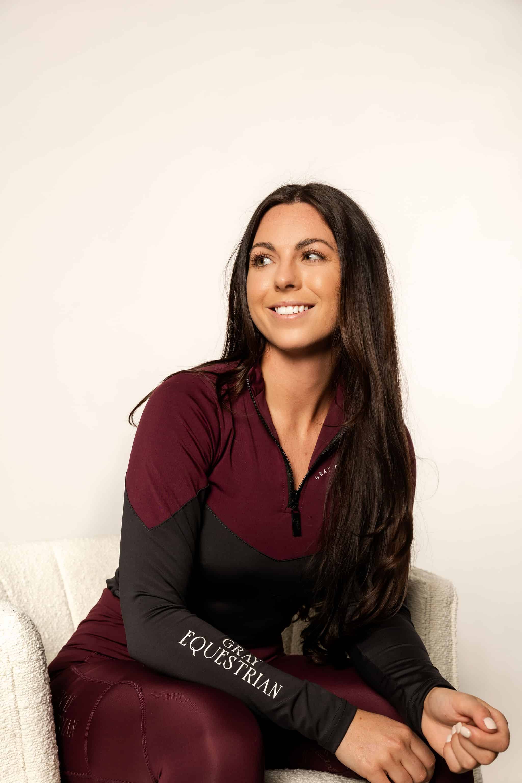 Brunette model wearing burgundy and grey base layer top with burgundy riding leggings in chair.