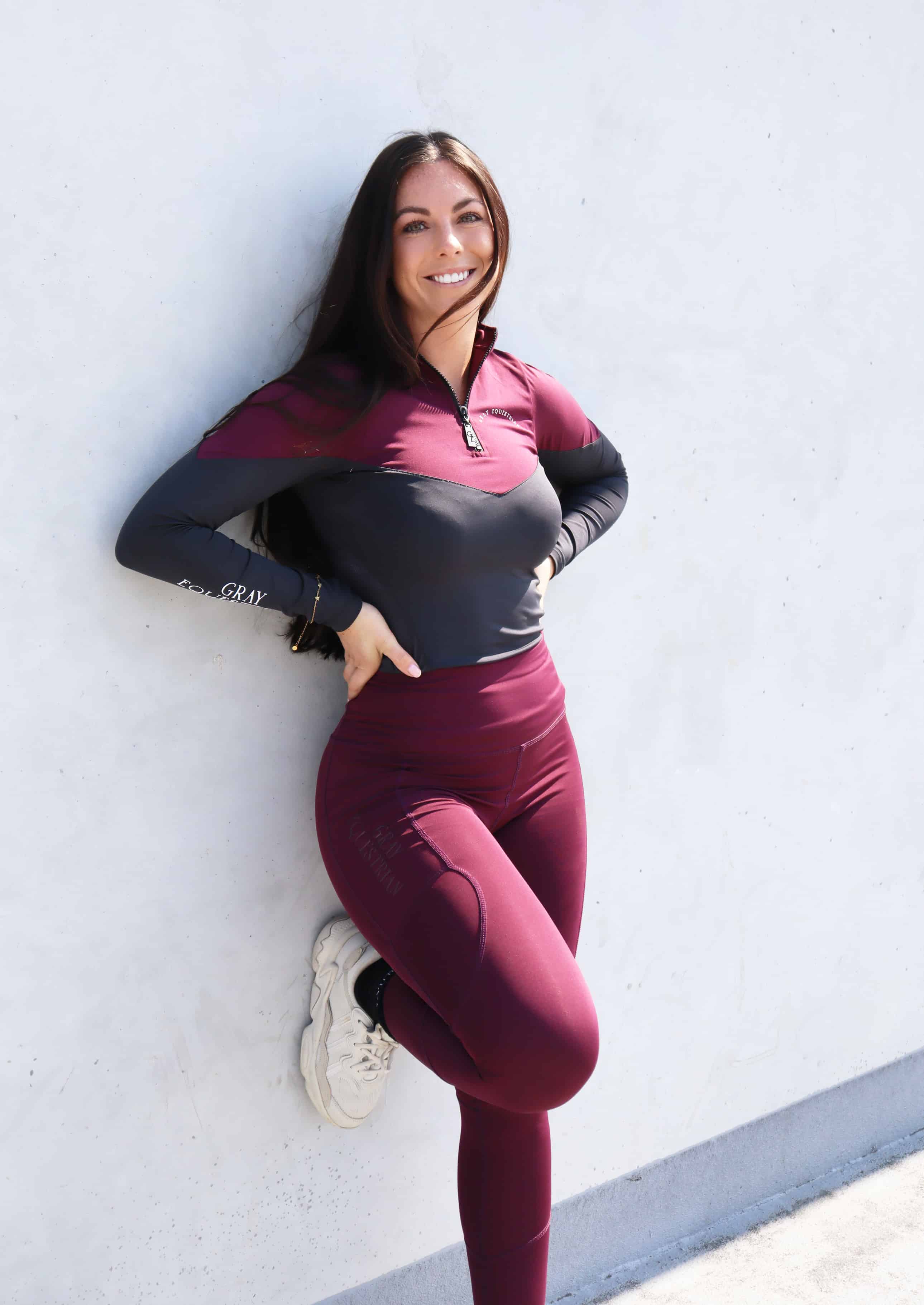 Brunette model wearing burgundy and grey base layer top with burgundy riding leggings leaning against a wall