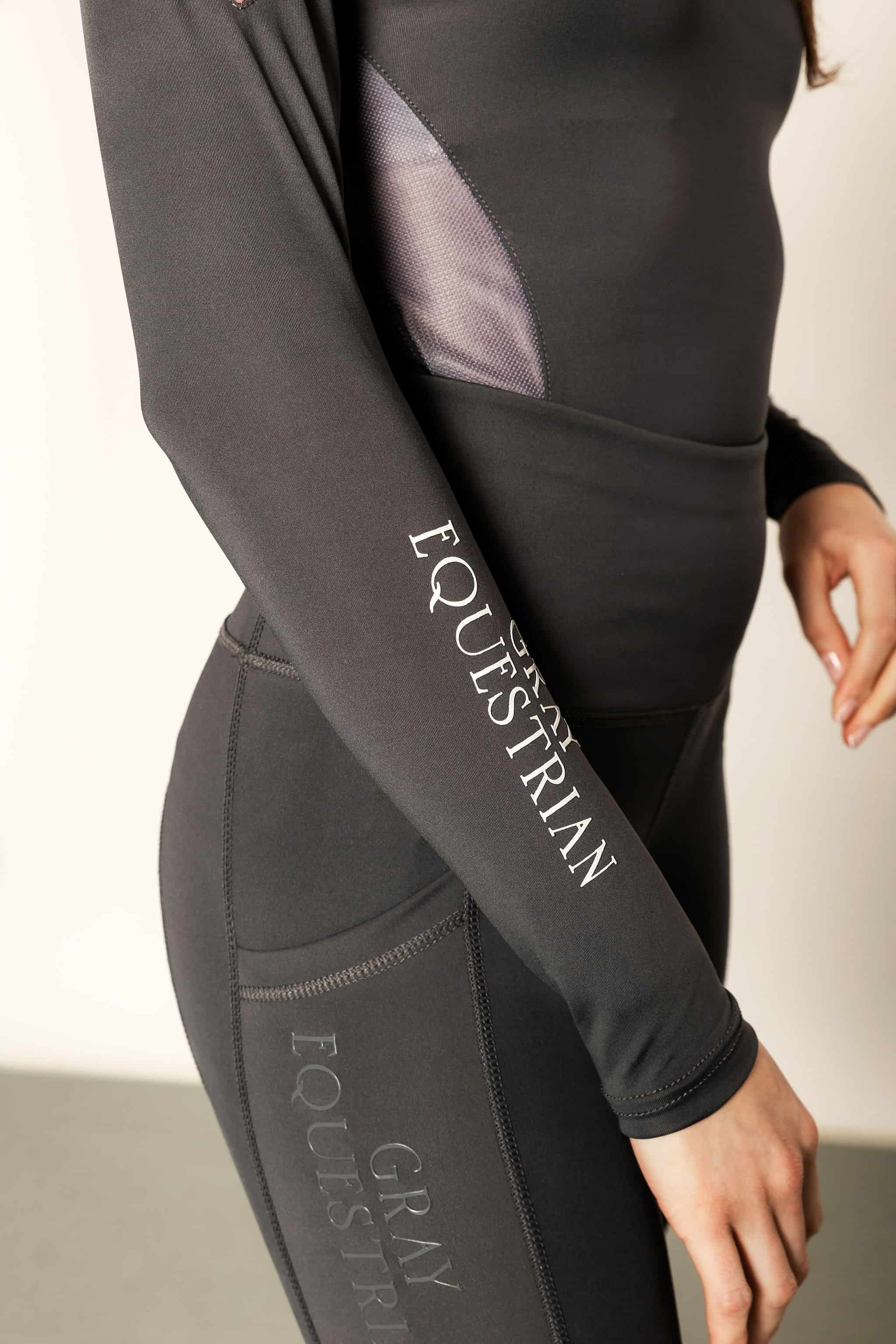 Model wearing charcoal grey riding leggings with pink and grey base layer top with white details.