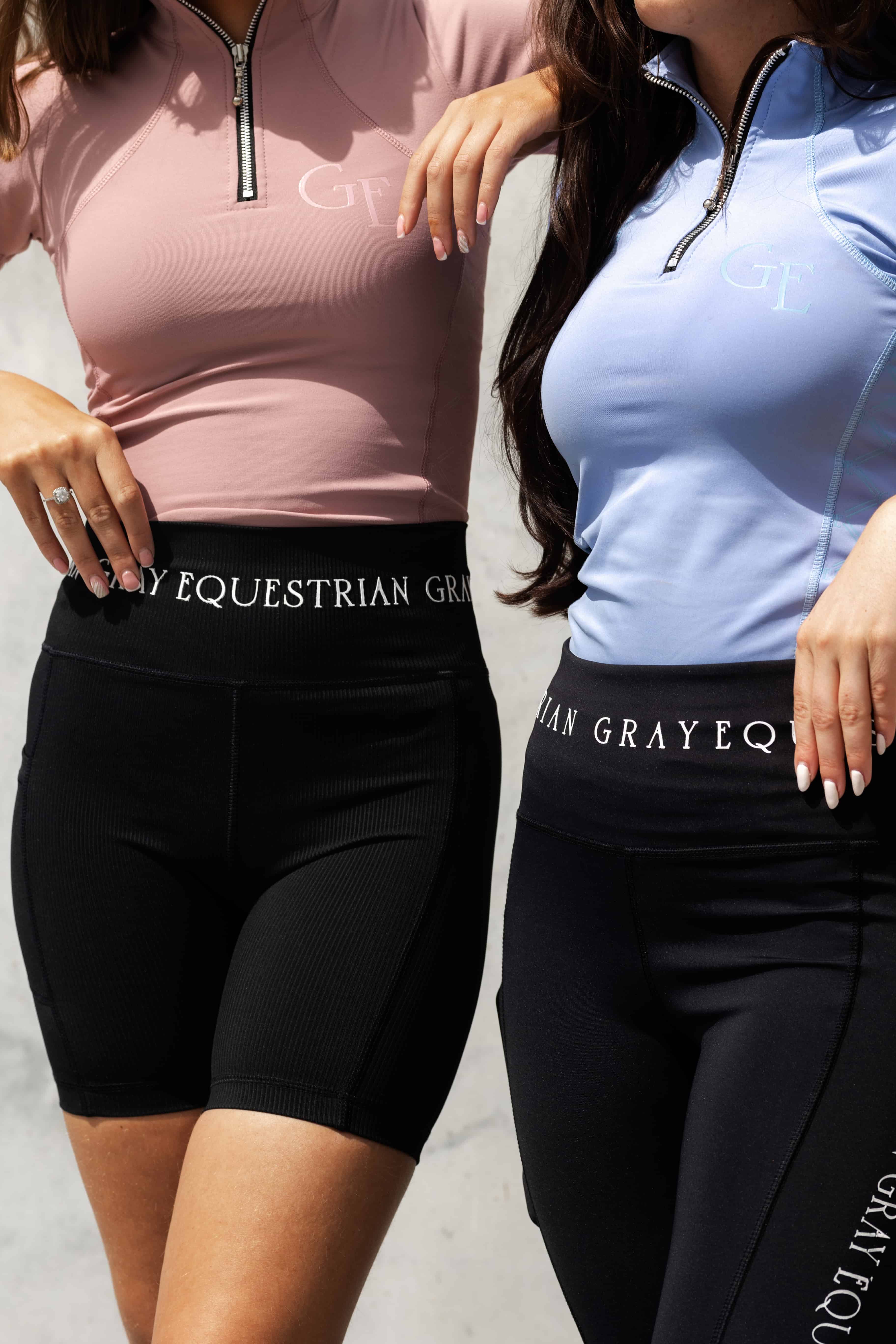 Two models sitting site by side. The model on the right is wearing our light blue base layer and black leggings. The model on the left is wearing our pink base layer and black riding shorts.