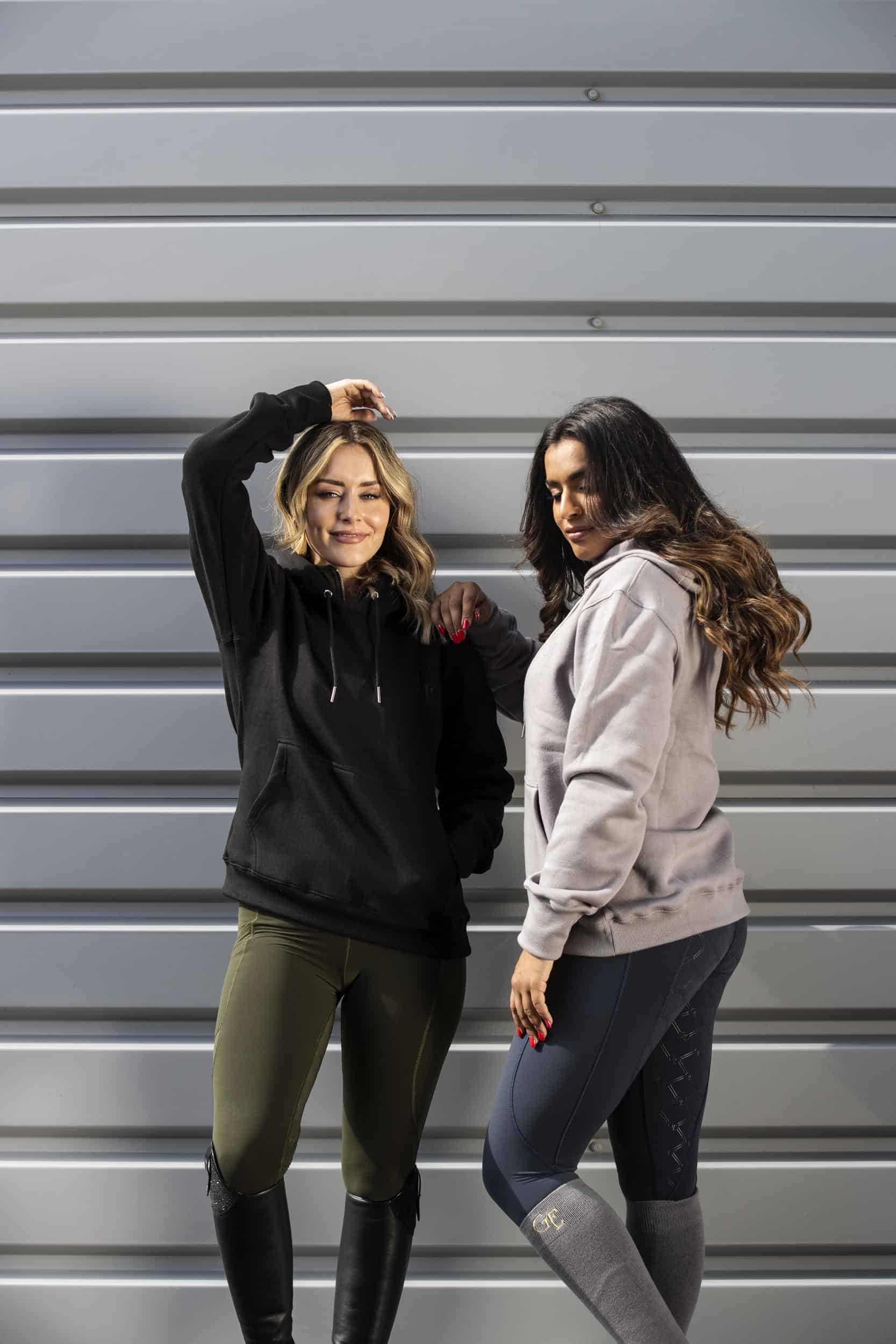 Two models standing next to each other. The model on the left is wearing our black hoodie and khaki riding shorts. The model on the right is wearing our taupe grey hoodie and navy riding leggings.