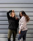 Two models standing next to each other. The model on the left is wearing our black hoodie and khaki riding shorts. The model on the right is wearing our taupe grey hoodie and navy riding leggings.